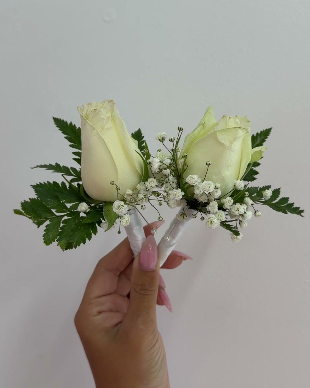 Two white rose and baby's breath buttonholes held against a white wall