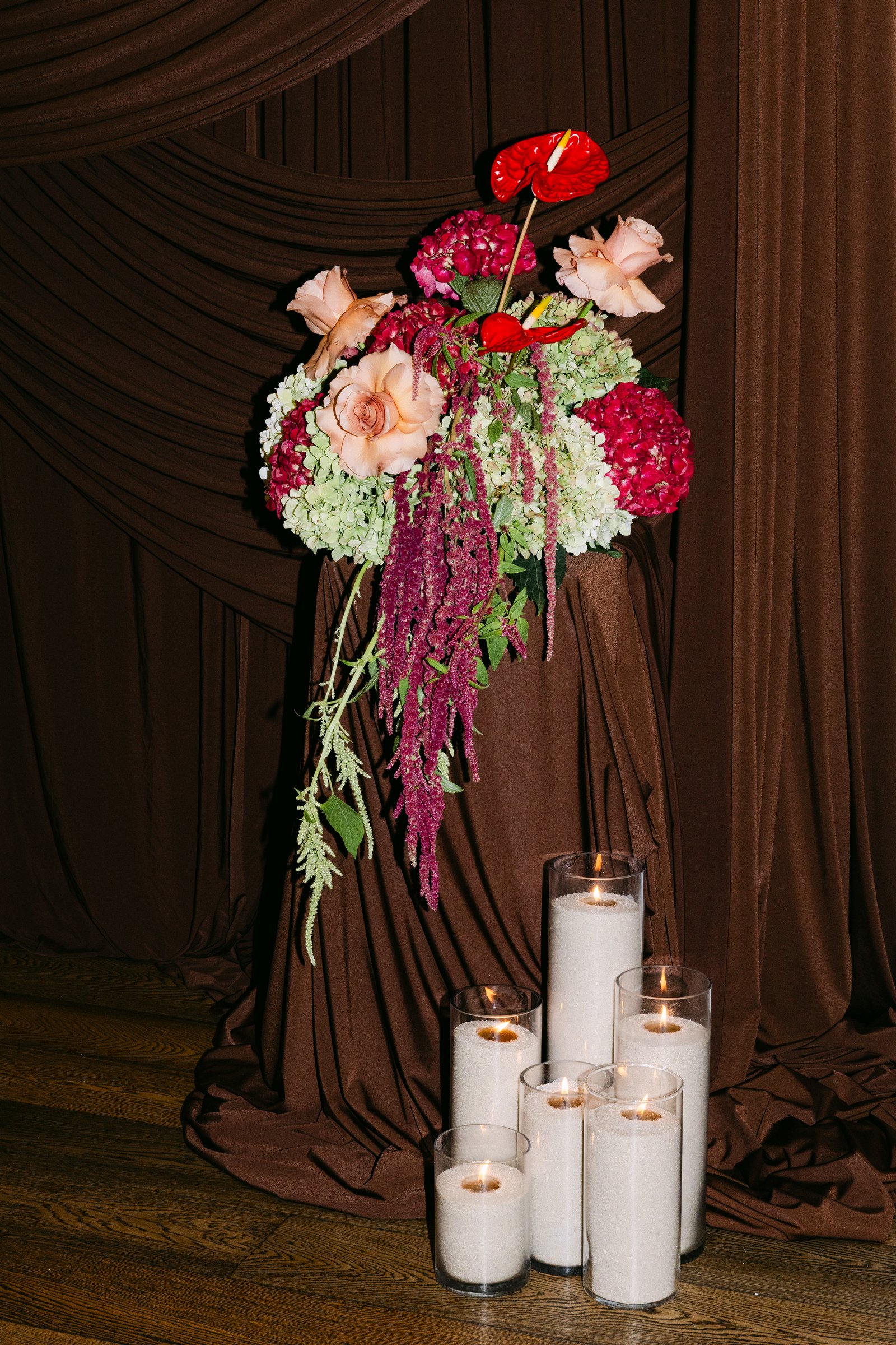 Red anthurium, hydrangea and trailing amaranthus arrangement on a draped plinth with pillar candles