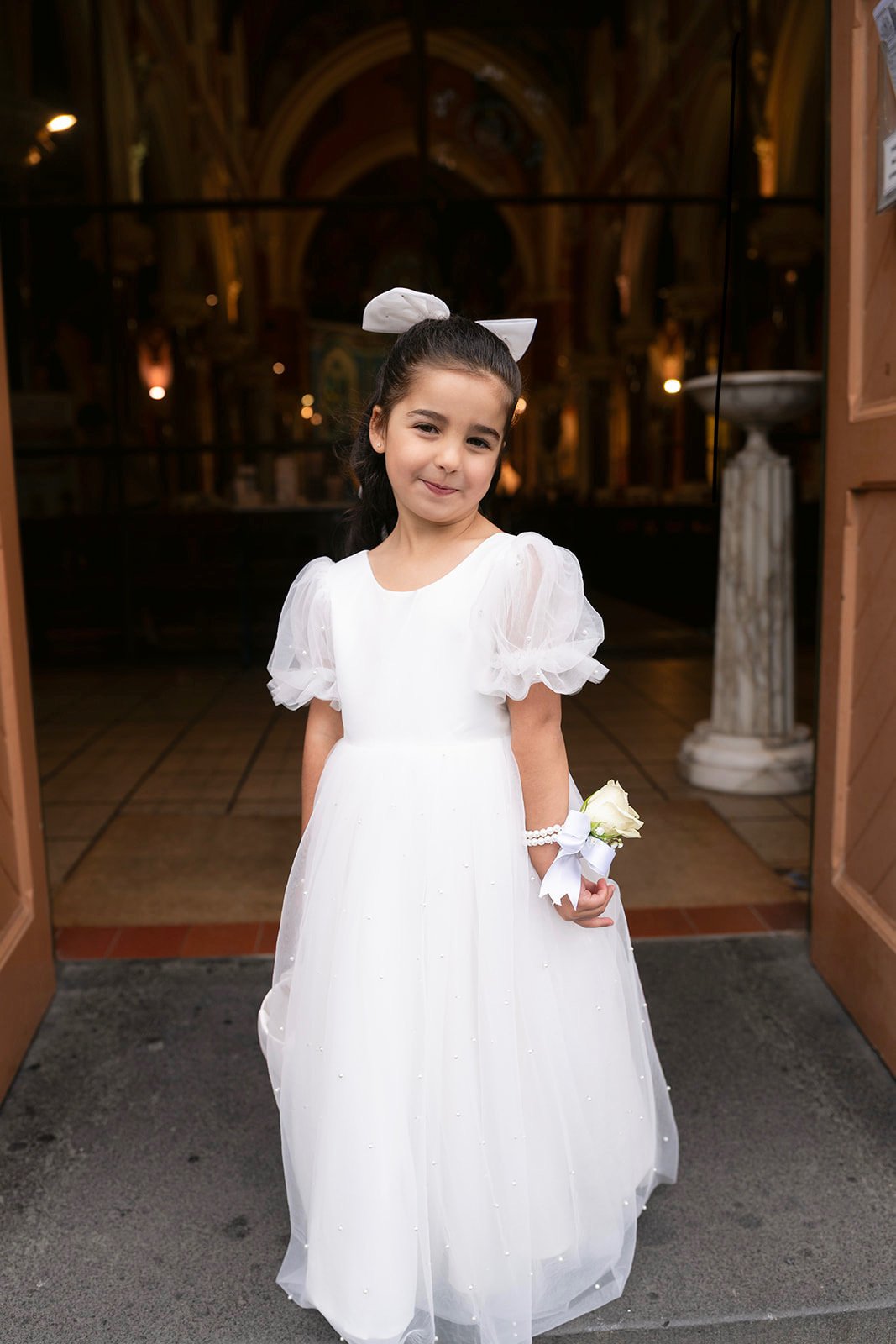 Flower girl in a white tulle dress with a rose wrist corsage at the church doors